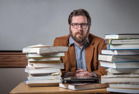 Paul Baltimore photo with stacks of books in front of him at teacher's desk